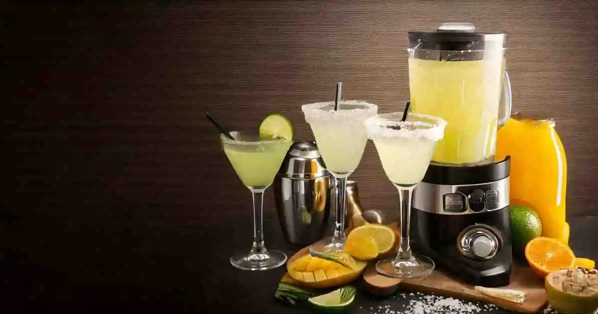 A setup featuring a Margaritaville machine with freshly blended margaritas in a pitcher and served in salt-rimmed glasses. The scene includes lime wedges, orange juice, sliced mango, and a cocktail shaker, all on a wooden board with a brown textured background.