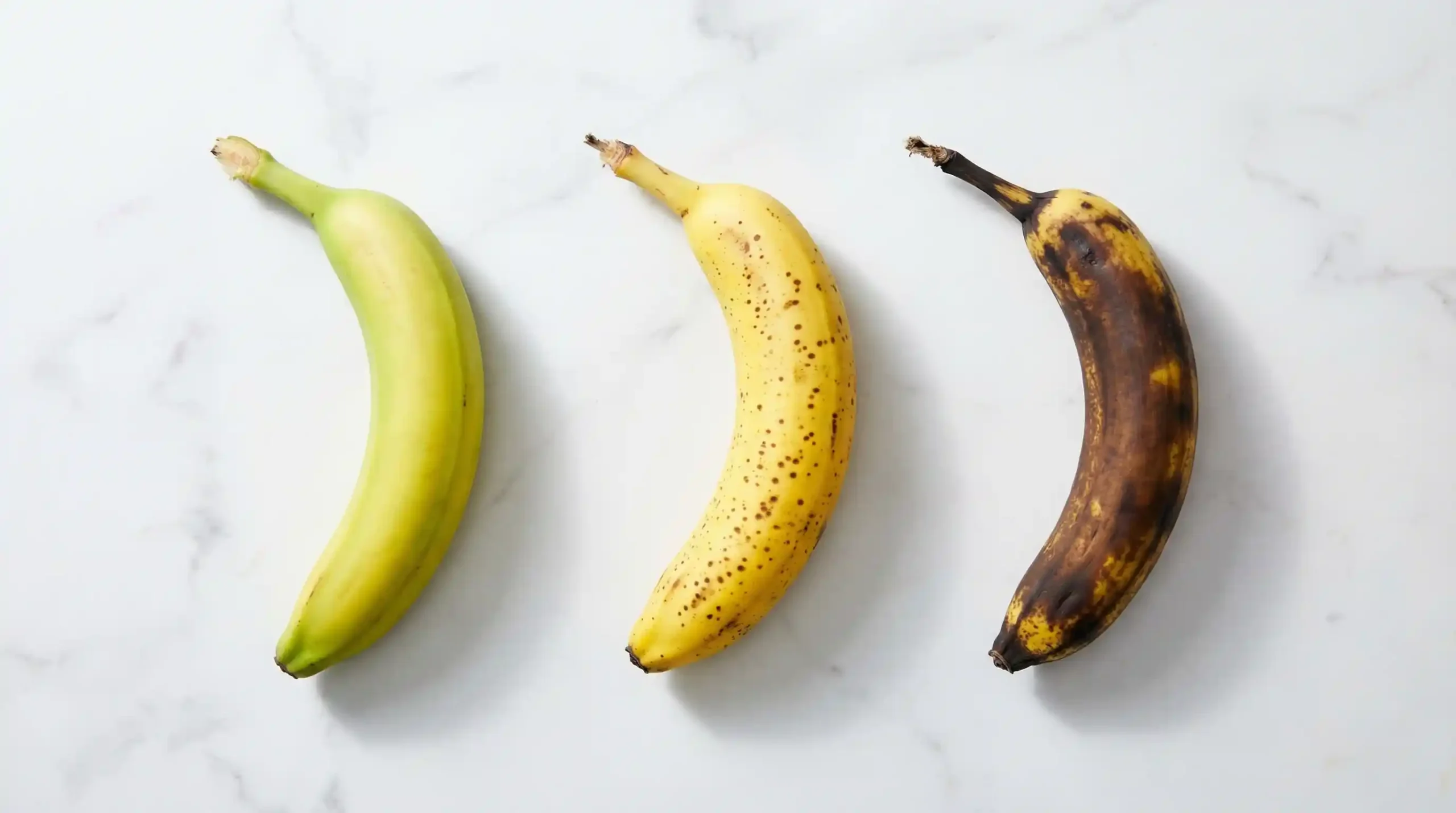 Top-down comparison of three bananas on a marble counter showing ripeness stages: green, yellow with spots, and brown overripe bananas perfect for baking.