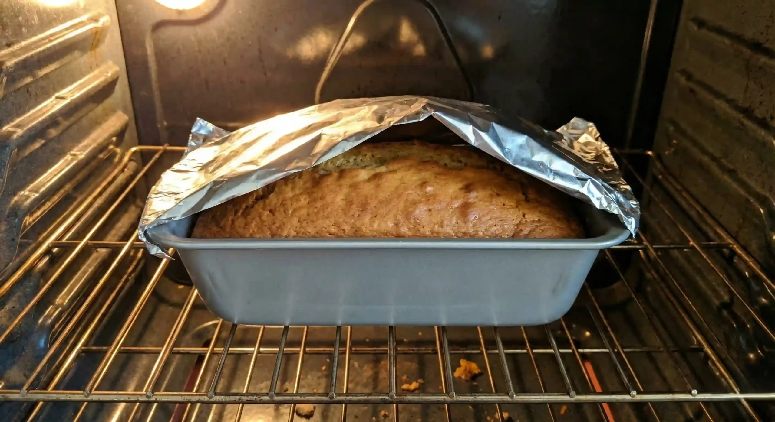 Banana bread loaf baking in an oven with aluminum foil loosely tented over the top to prevent the crust from burning while the center finishes cooking.